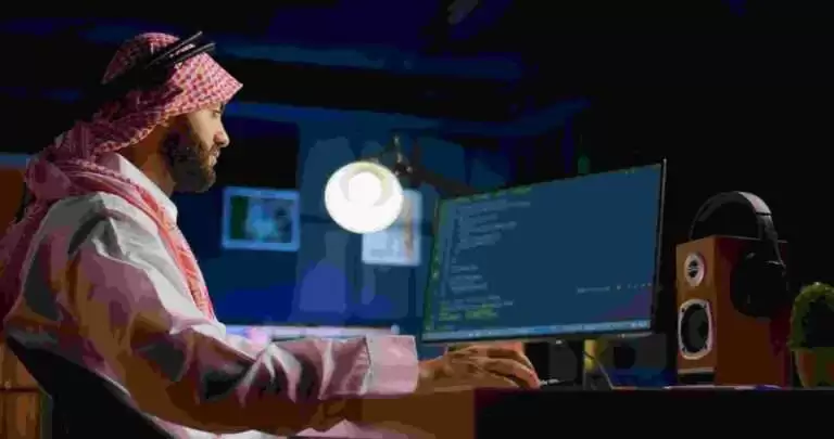 A man wearing traditional Middle Eastern attire works on a computer, analyzing code for a cybersecurity company in Dubai. He is seated at a desk in a dimly lit room with a large monitor displaying programming scripts. A pair of headphones rests on a wooden speaker beside him, and a small potted plant is visible on the desk.